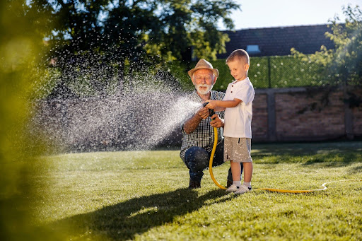 “手入れが行き届いた青々とした芝生の上で、笑顔で水やりを楽しむ祖父と孫の姿"